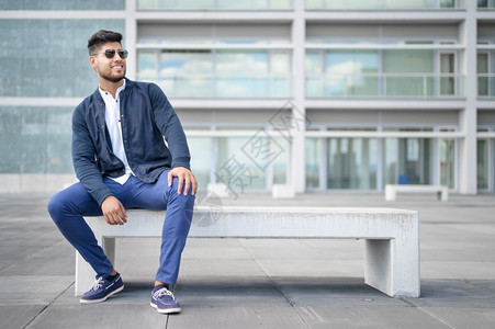 身着时尚太阳镜的年轻男子肖像在城市背景中坐长椅上摆姿势身着时装太阳镜的年轻男子画像在城市背景中坐长凳上摆姿势夹克全套服装现代的图片