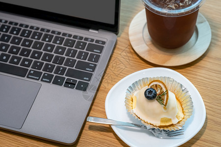 在咖啡馆店的木制办公桌上在商业工作期间用键盘笔记本电脑在咖啡厅的木制办公桌上关闭咖啡杯中的冰和自制白橙蛋糕面包店木制的休息图片