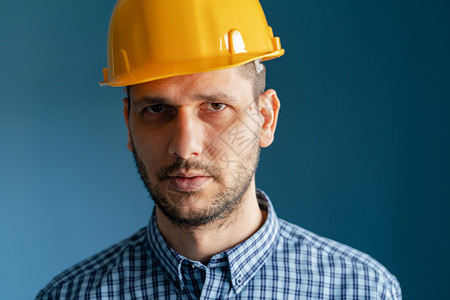 工人建筑师在蓝墙前穿着黄色保护安全头盔和衬衫的caucasian年轻男子工程师近距离肖像观看摄机的面部被隔绝安全的图片