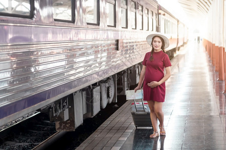 在火车站旅行时身着红裙子的亚洲孕妇持有一张带红色行李箱的在火车站旅行时身穿红色衣服的亚洲妇女在夏季假期概念泰国旅行者携带背包手淑图片
