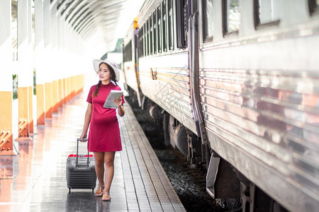 女白种人在火车站旅行时身着红裙子的亚洲孕妇持有一张带红色行李箱的在火车站旅行时身穿红色衣服的亚洲妇女在夏季假期概念泰国旅行者携带图片