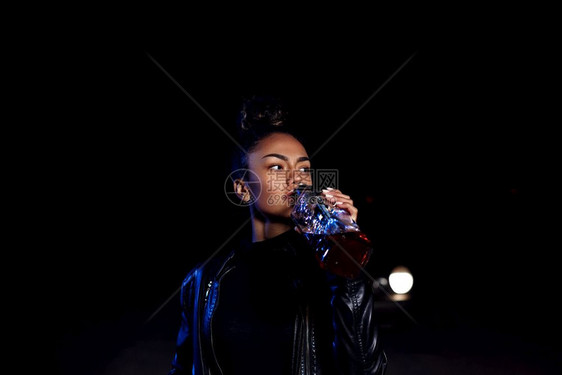 一种生气的走身穿皮夹克和黑衣服的非洲醉酒年轻黑人穆拉托女孩在一条废弃沙路上在汽车灯光和街道照亮下夜里喝着一瓶威士忌图片