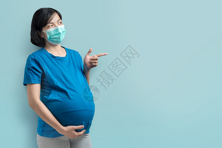 福利穿着保护面罩和蓝色T恤衫指着并寻找空间的笑快乐亚洲女孕妇她们戴着保护面罩和蓝色T恤试图模仿空间女站在孤立的背景下触摸肚子医疗图片