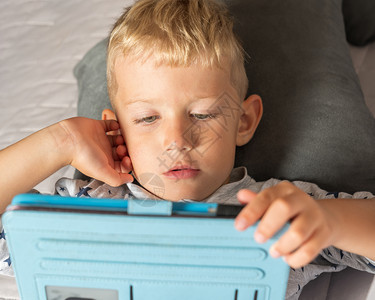 享受正面无线的白种男孩在家里躺床上时使用数字平板电脑图片