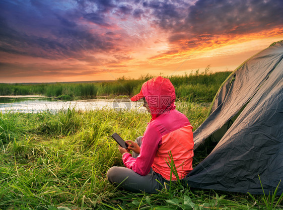 女孩靠近水穿粉红色夹克的妇女与手机坐在帐篷附近一位女士与河岸上一个帐篷在戏剧般的天空下夏月风景的旅行和自由概念穿粉红色夹克的妇女图片