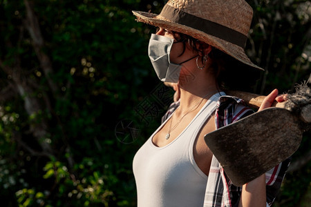 年轻农民妇女带着草帽走动的外科面具和蹄子望着照相机工人背心新冠图片