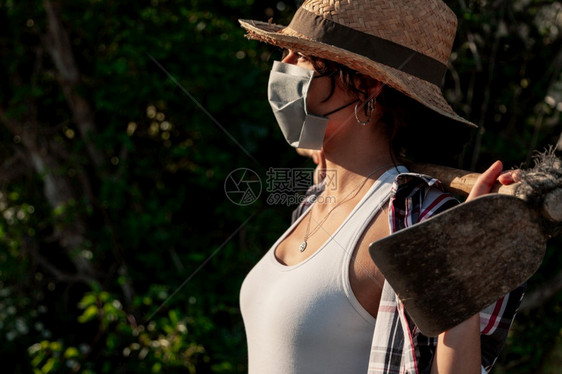 年轻农民妇女带着草帽走动的外科面具和蹄子望着照相机工人背心新冠图片