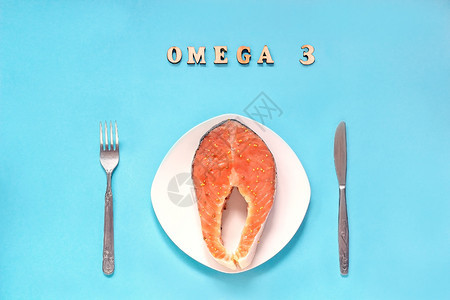 白盘刀叉上的红腌鱼牛排片蓝色背景上的静物和铭文Omega3平躺顶视图白盘刀叉上的红腌鱼牛排片盘子新鲜的健康图片