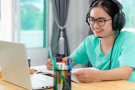 快乐的亚洲人青年女学生带着眼镜耳机的亚洲女青年学生在一本书上寻找视频会议笔记本电脑计算机大学课程在线网上习络教育在家中学习远程教图片