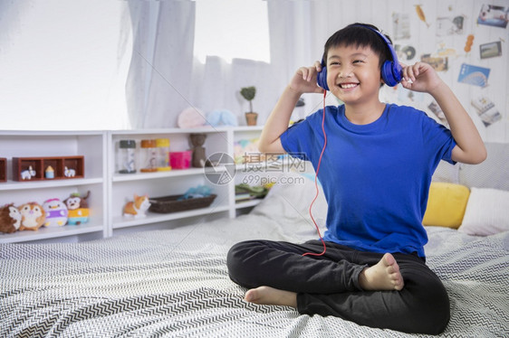 在室内歌曲蓝耳机的亚洲男孩使用智能手机在家里床上听音乐时笑着快微在家中听到音乐白种人图片