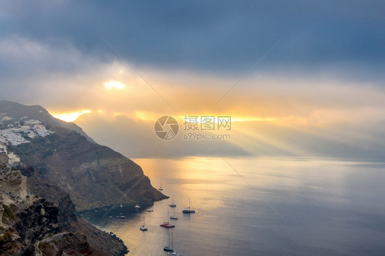 希腊桑托里尼太阳射线卡路德拉上空的重云照亮了海湾和数艘停靠的游艇圣托里尼卡尔德拉和穿透重云的太阳射线日出悬崖景观图片