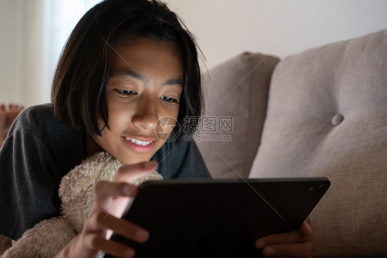 近距离拍摄的亚洲小女孩使用平板电脑在沙发上躺观看录像中笑着微和在家中脸上表情房间学习在线的图片