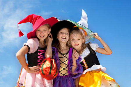 秋天巫婆阳光三个美丽的微笑小女孩长发在巫服装中看着镜头在蓝天空A用诡计或处理袋图片