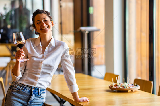 一位年轻美丽的黑人女子肖像她站在桌边靠家中举着一杯红酒独自微笑着穿白色衬衫看镜头前视线微笑着容酒精真实的白色图片