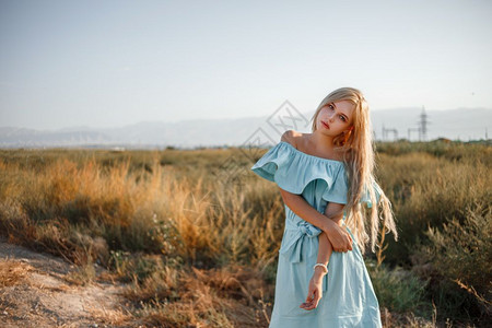 一种乡村的身着浅蓝色轻礼服站在一个田地上旁边一条小乡村路旁有晒太阳草的年轻美丽caucasian金发美女肖像模糊图片