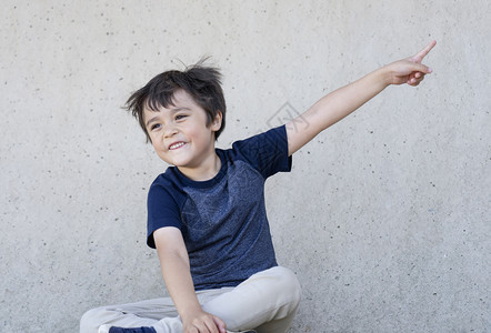 享受快乐的小男孩肖像带着微笑的脸举起兴奋的小孩脸坐在灰墙旁边用指着手孩子在户外玩得开心积极的儿童概念男生吸引人的图片