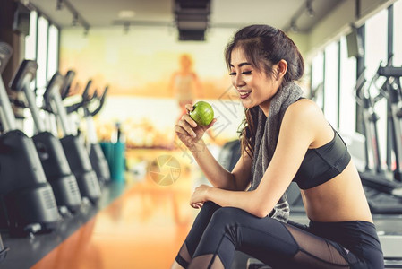 身体吸引人的燃烧亚洲女持有并寻找绿色苹果与体育设备和跑步车一起在背景中用运动设备吃饭清洁食品和健康概念健康身锻炼运行主题图片
