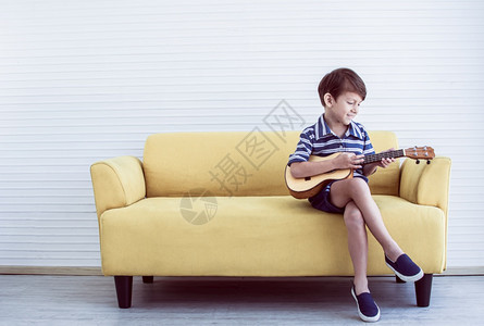 俏皮在家中客厅的黄色沙发上弹吉他时Caucasians男孩的肖像很快乐玩初学者图片