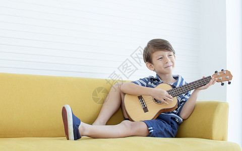音乐活的唱歌在家中客厅的黄色沙发上弹吉他时Caucasians男孩的肖像很快乐图片
