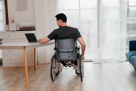 车轮在室内桌子坐轮椅上家用电脑工作的快乐亚洲残疾人男子在家中用电脑工作图片