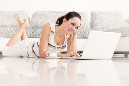 家美丽的年轻女在笔记本电脑饮用水咖啡客厅地板上躺着的互联网冲浪人们诺维科夫图片