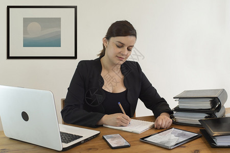 基多坐着美丽的西班牙裔女行政办公室职员笑得开心坐在办公桌前工作忙着在笔记本上写字靠在白墙上笔记本电脑图片