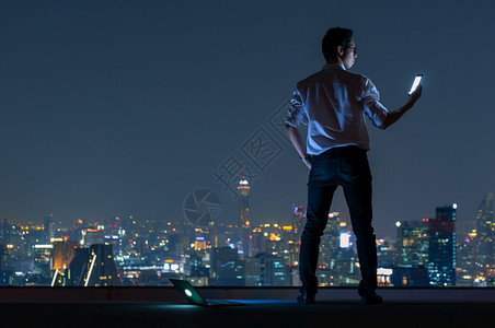 套装亚洲商人站在大楼顶端使用智能手机和笔记本电脑夜视城市的风景商业成功与技术概念在深夜工作利用智能手机和笔记本电脑在夜间观察城市图片
