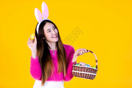 亚洲人庆祝穿着兔子耳朵的亚洲快乐女青年手拿着一个篮子配彩色复活节鸡蛋在黄色空版间工作室背景上孤立无闻季节图片