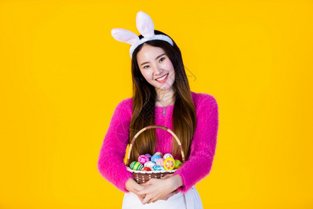 假期美丽的季节穿着兔子耳朵的亚洲快乐女青年手拿着一个篮子配彩色复活节鸡蛋在黄色空版间工作室背景上孤立无闻图片
