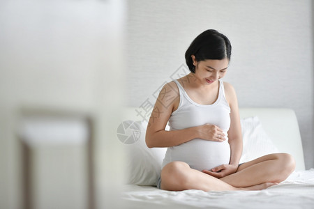 愉快腹部怀着白帕贾马斯的美丽亚洲孕妇身穿白色睡袍坐在床上微笑着健康快乐的年轻母亲在怀孕时抱着摸和看肚子在家放松图片