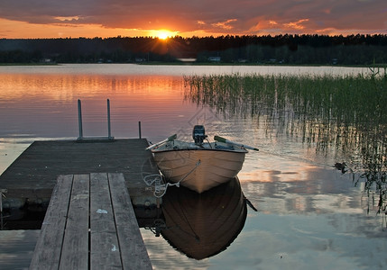 木制的斯堪纳维亚树木在和平的多彩日落时由木制小船停靠在日落时被木制小船停靠在码头瑞典瓦尔姆兰一个湖上以宁静的水中天空反射着图片