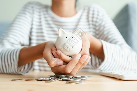 利润支付亚洲年轻女孩手握着白猪银行的亚洲年轻女孩每天早晨在家里用两只手硬币堆上退休金融和为未来概念储蓄资金小猪图片