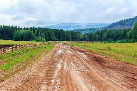坏的俄罗斯西伯利亚乡村地带绿和木林山之间的地平线上以图象湿润的泥土道路为景色的夏季风碎石字段图片