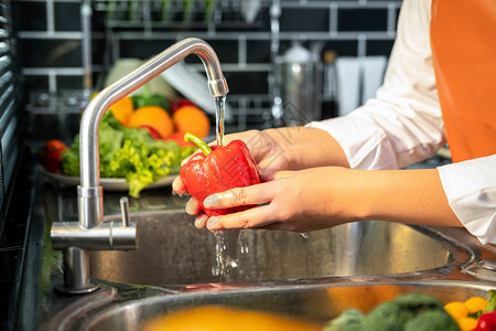 亚洲家庭主妇穿围裙站着洗胡椒和蔬菜在家中厨房做饭以获得健康餐时用水槽中浇卫生为了美食图片