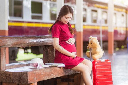 亚裔怀孕妇女穿着红色礼服坐在长椅上背着红行李乘火车站旅行的TeddyBear和TeddyBear女孩沟通幸福图片