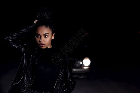 身着皮夹克和黑衣服的非洲裔美国女孩穆拉托Mulatto在一辆废弃沙路上晚在车灯和光照亮下时髦女摆姿势图片