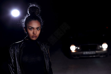 脸化妆品皮革身着夹克和黑衣服的非洲裔美国女孩穆拉托Mulatto在一辆废弃沙路上晚在车灯和光照亮下图片
