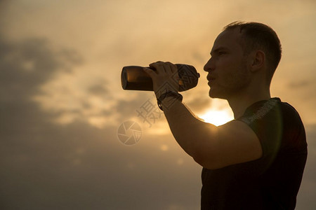 慢跑者男人晚上身穿黑色T恤和短裤的白人体育青年男子在日落时喝瓶酒饮用水的肖像图片