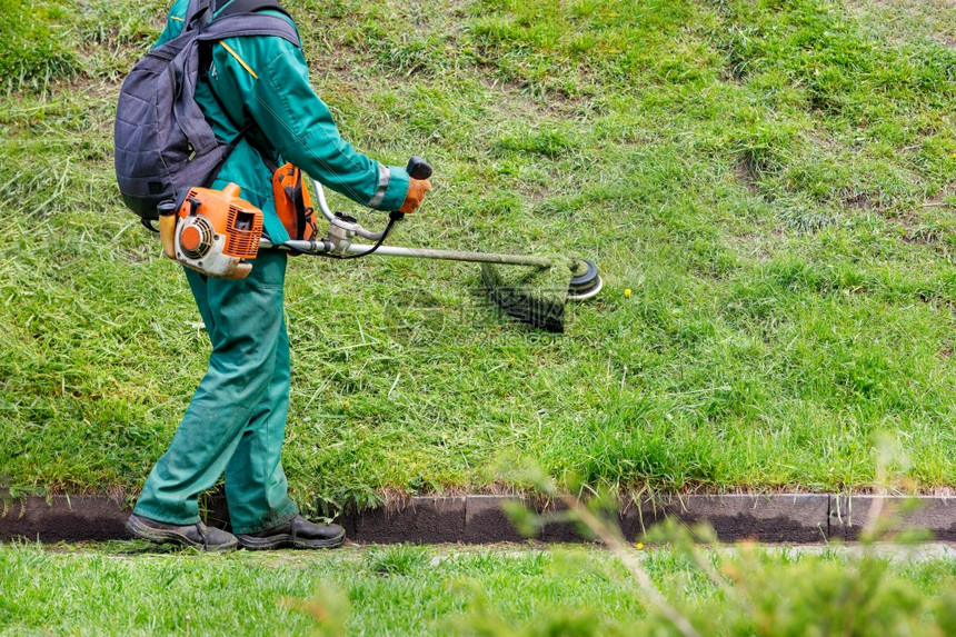 环境一名身穿绿衣的工人用一个业汽油在城市公园里使用一个工业汽油加分器来割草A工人用汽油加分器在山上撒绿草发动机切割图片