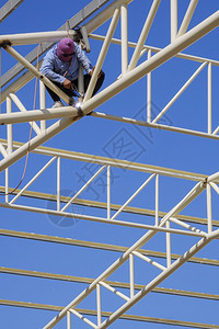 眼镜亚洲建筑工人的低角度观点亚洲建筑工人在垂直框架蓝天空背景下将金属焊接在屋顶建筑结构上并在垂直框中结合蓝色天空背景垂直的低图片