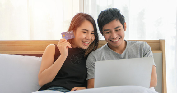 买亚洲夫妇在线购物用笔记本电脑信卡付款在家床上冲网时快乐的一对夫妇保持买方图片