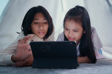 可爱的人们两个快乐亚裔女孩晚上在帐篷里用平板在帐篷里使用亚洲儿童拥抱娃在家数字电脑上学习愉快图片