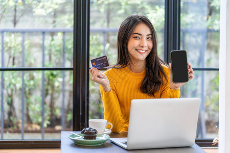 使用信卡的亚洲年轻妇女在咖啡店或合用空间咖啡杯技术资金钱包和网上付款概念信用卡模型上展示移动电话在线购物时可以使用信卡和手机进行图片