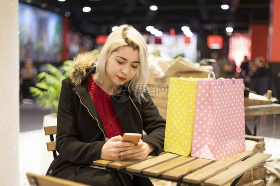 店铺客户年轻的提着购物袋年轻女子坐在购物中心的咖啡馆里手拿着电话的女孩购买太多概念快乐购物提着袋的年轻女子坐在购物中心的咖啡馆里图片