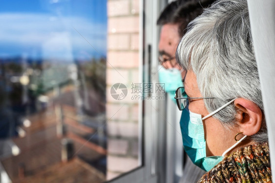 戴防护面罩的老年夫妇在家看窗社交距离封锁老人和退休的生活方式常设面具关心图片