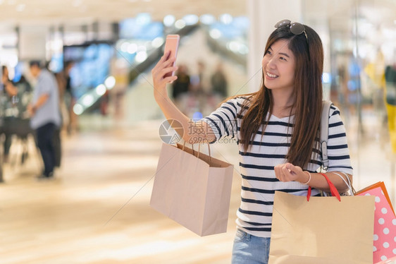 购物曼谷折扣亚洲妇女步行使用智能手机自拍模特通过商店面的眼镜展示装饰服的模型在部门中心欢乐地行动起来亚洲妇女走路用智能移动电话自图片
