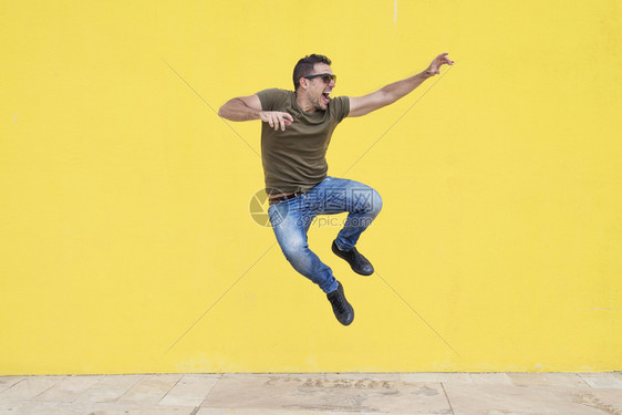 全套服装带着太阳眼镜的年轻人在黄墙自由前跳跃着垂直的活力图片