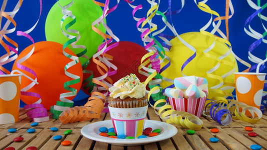 甜点土生木制餐桌上的杯子蛋糕背景是彩色气球塑料杯和带蓝墙的糖果乡村纸杯蛋糕图片