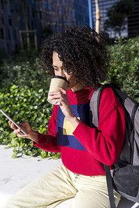 杯子坐在户外长椅上喝咖啡杯时使用手机的美丽年轻女青热的毛衣图片