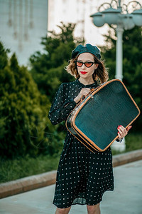 喜悦周末复古的身着老黑色波尔卡圆点装饰的年轻女子手持复式提箱在外面摆放图片